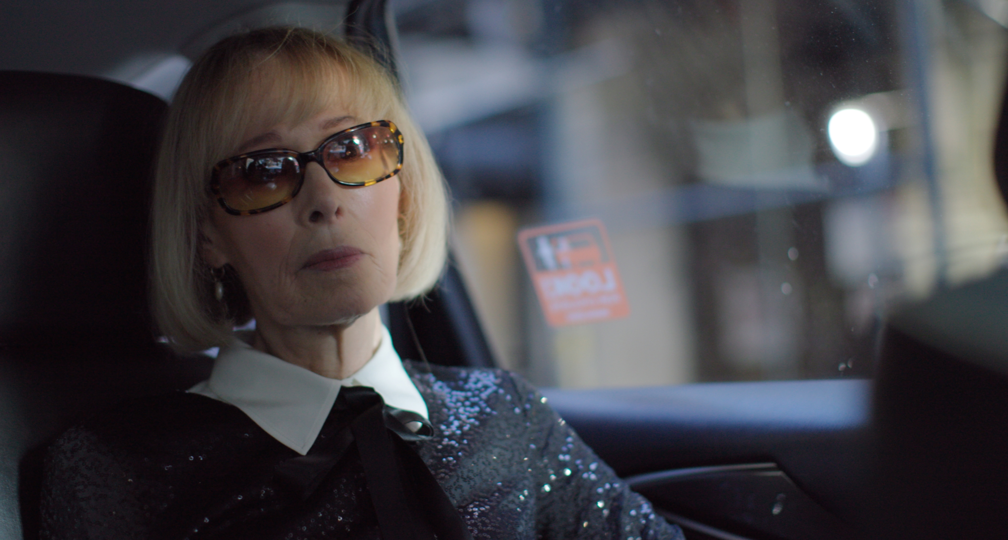 writer e. jean rides in the back of a car wearing sunglasses and a sparkly buttoned up top in a still from the documentary ask e jean