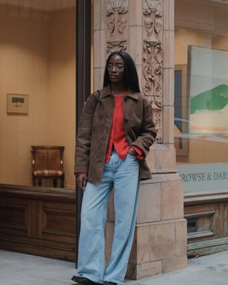 London woman wears light wash baggy jeans with a red cardigan, brown suede jacket, and loafers.