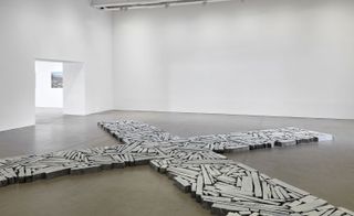 A gallery space with a large cross laying on the grey floor make from pieces of concrete