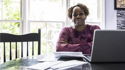 A satisfied-looking woman sits in front of her laptop at the kitchen table.