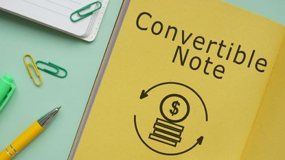 The words "convertible note" on a yellow notebook next to some desk supplies.