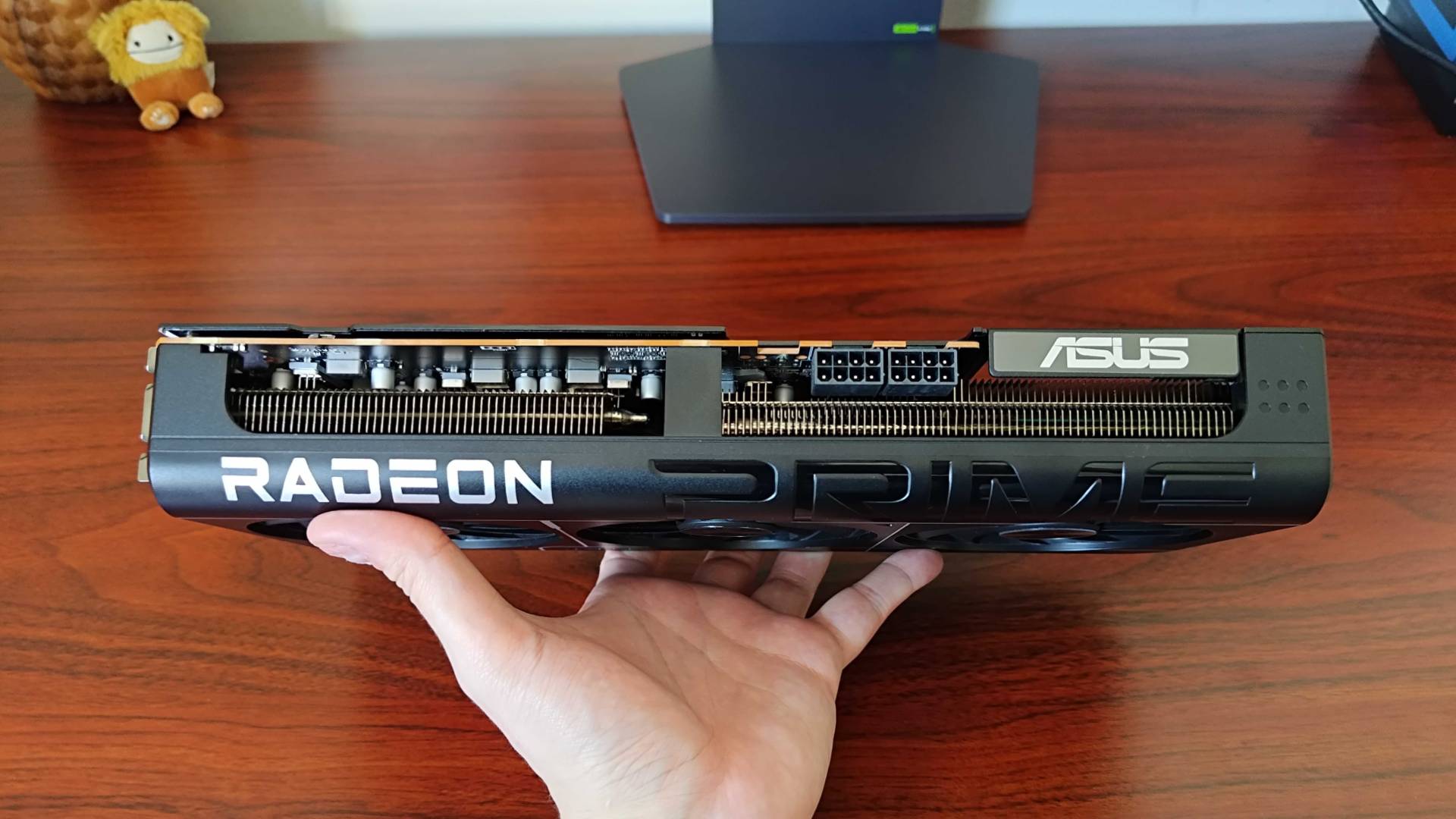 Hand holding AMD Radeon RX 9070 Asus Prime graphics card next to woodgrain desk.