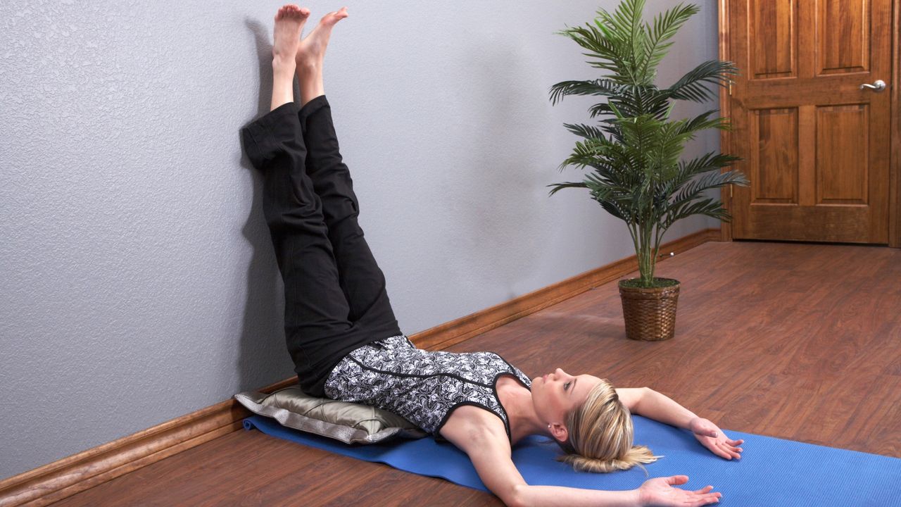 woman lying on her back with her legs raised up the wall. she&#039;s lying on a blue yoga mat on a wooden floor with a door and plant behind her. 