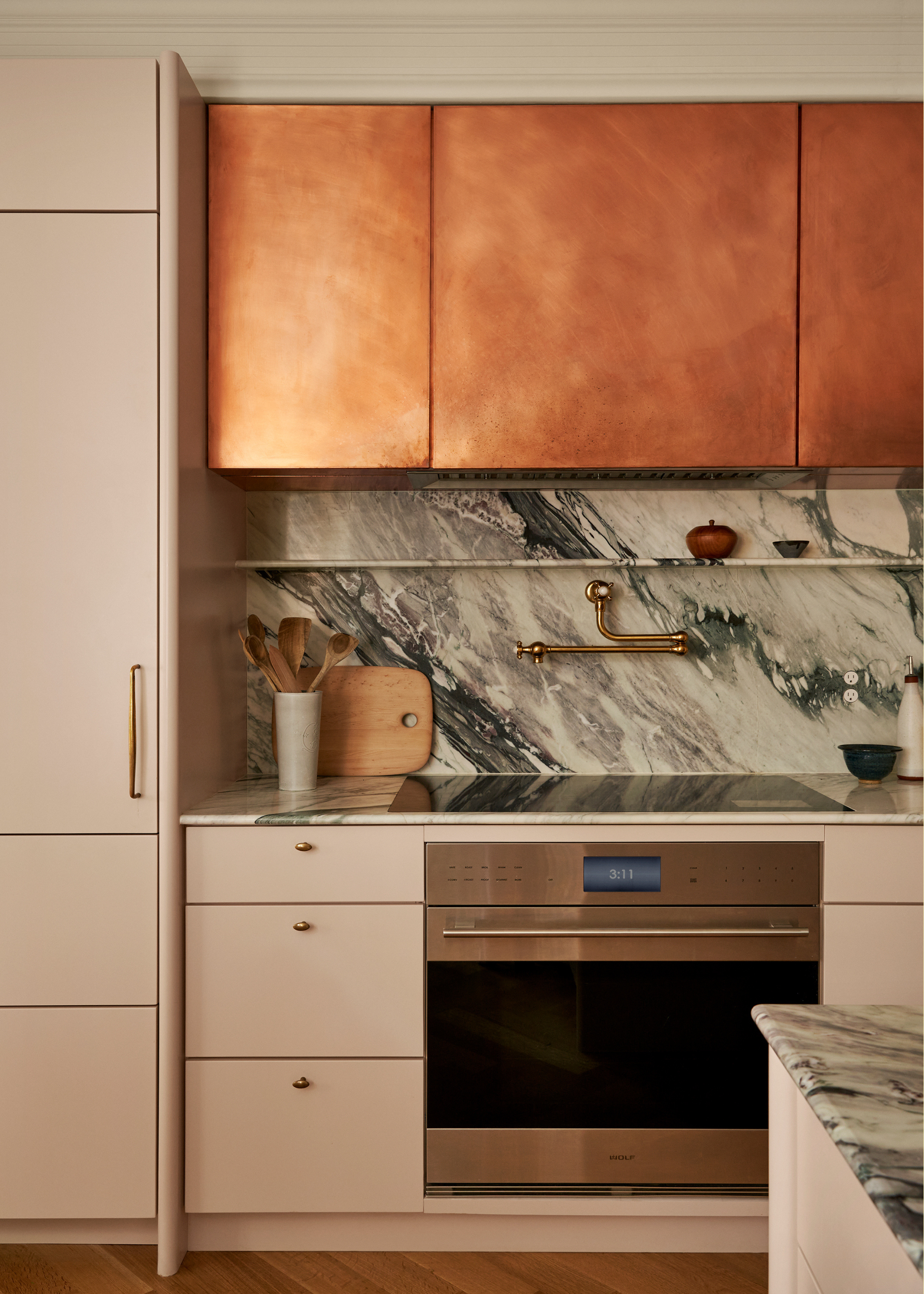 A beautiful kitchen with a stone counter and backsplash, brushed bronze cabinets, pale blush drawers, a hob, an oven, an extendable tap, and a chopping board by a vase of wood spoons