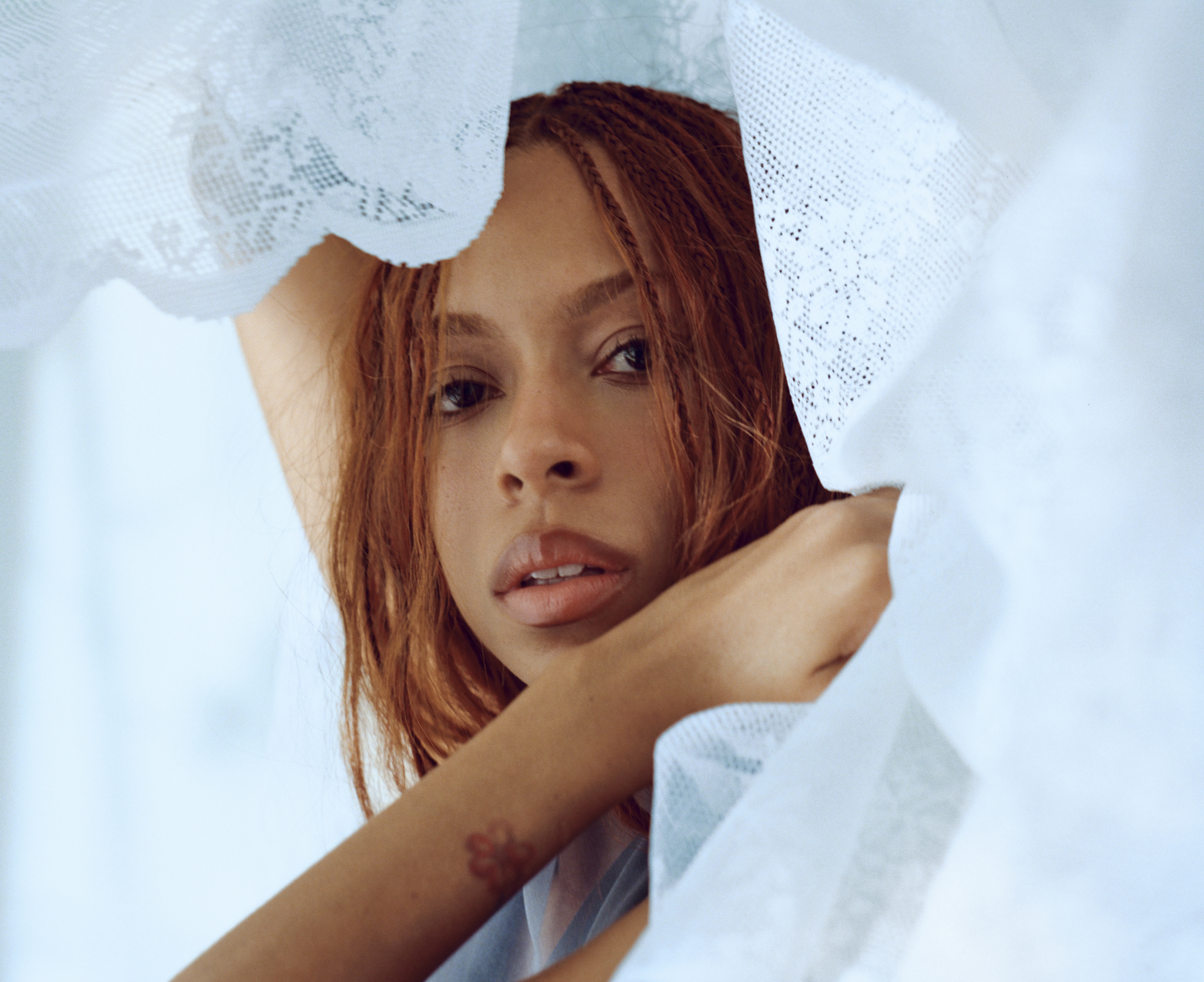 singer ravyn lenae looks at the cambera while posing in lace white sheets in a promotional shot for her album blue island