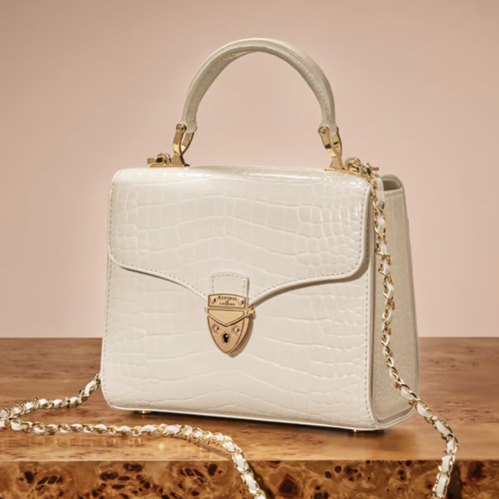 A white Croc-style Mayfair Aspinal of London crossbody bag on brown marble. 