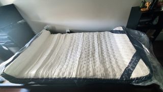 The DreamCloud Mattress shown unrolled but still vacuum packed in plastic and placed on our lead reviewer's bed frame