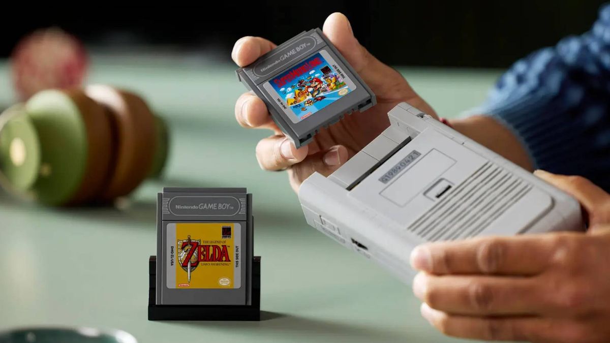 Hand inserting Lego Game Boy cartridge into model with Zelda cart on desk in backdrop.