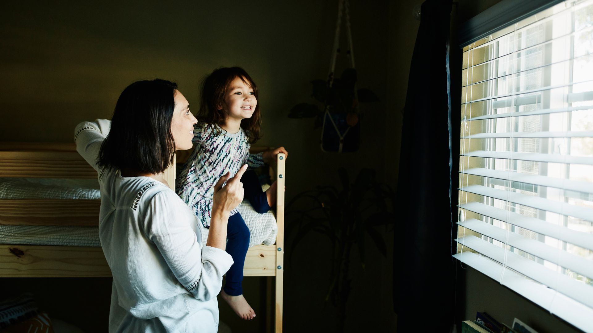 A mom and young child, who is sitting on the top bunk of bunkbeds, look out the window together in the morning.