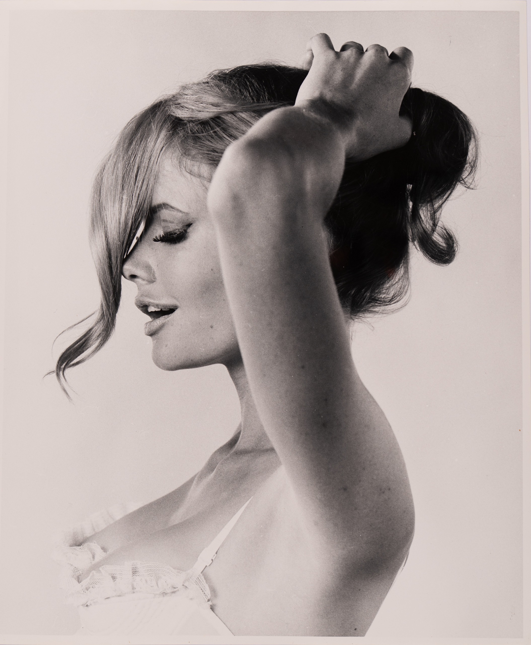 A woman in profile lifts her blonde hair with one raised arm, eyes closed and lips slightly parted, wearing a lace-trimmed top.