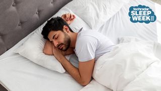 A man with dark hair and a beard sleeps on his side in a white bed