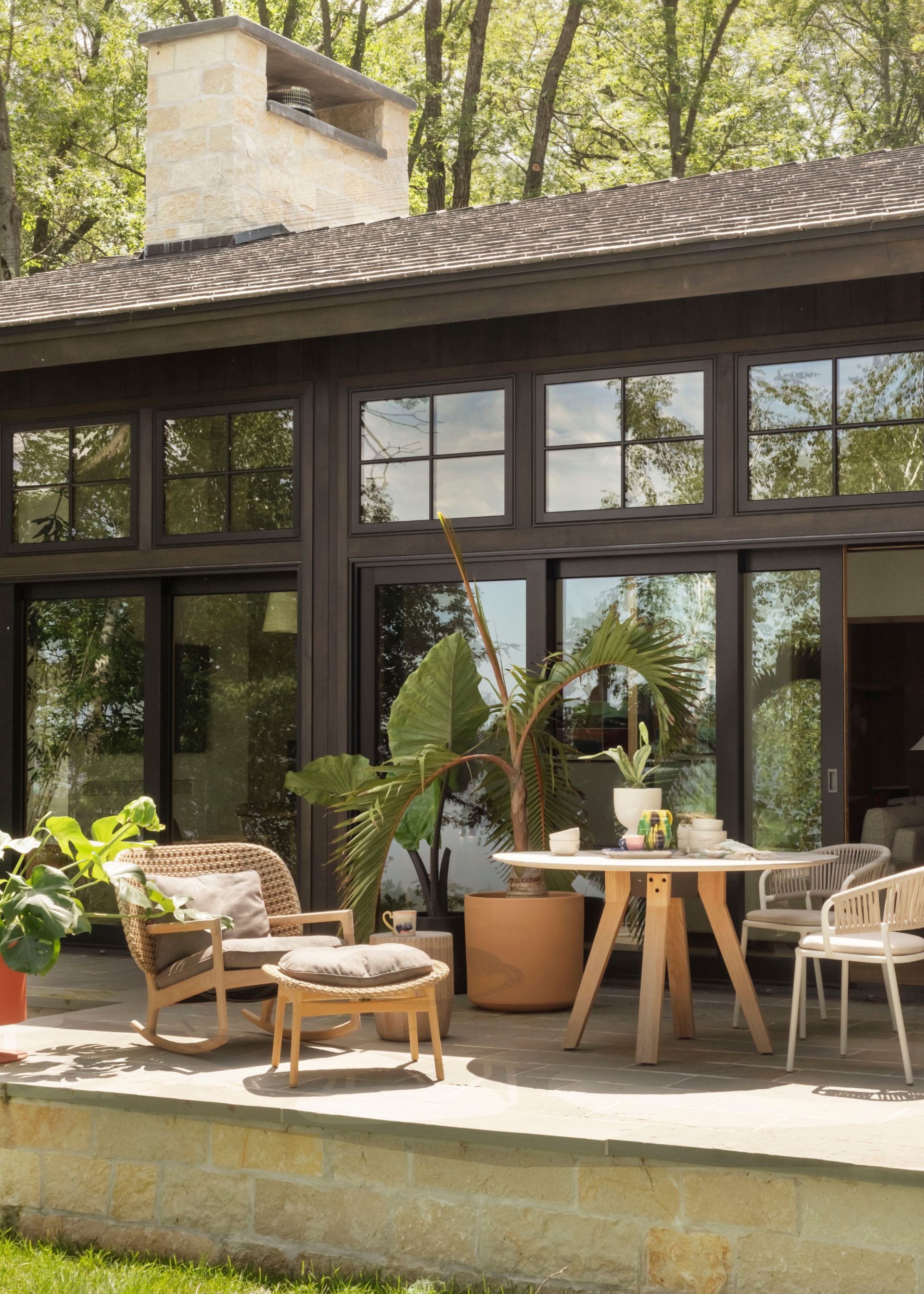 A calming patio area with a large potted plant next to an outdoor table and seating in warm, natural materials against a dark-wood home with sliding glass doors