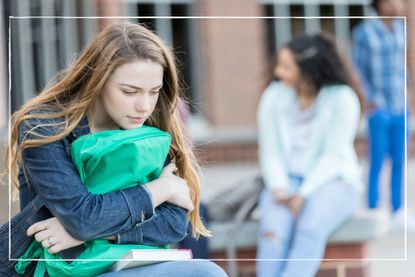 A teenage girl sits hugging her school bag, looking anxious