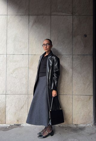 A cute work winter outfit is shown in a photo of a woman standing outside wearing a leather trench coat over a black knit polo styled with a gray wool maxi skirt, black tights, black eyeglasses, black mary janes and a black frame handbag