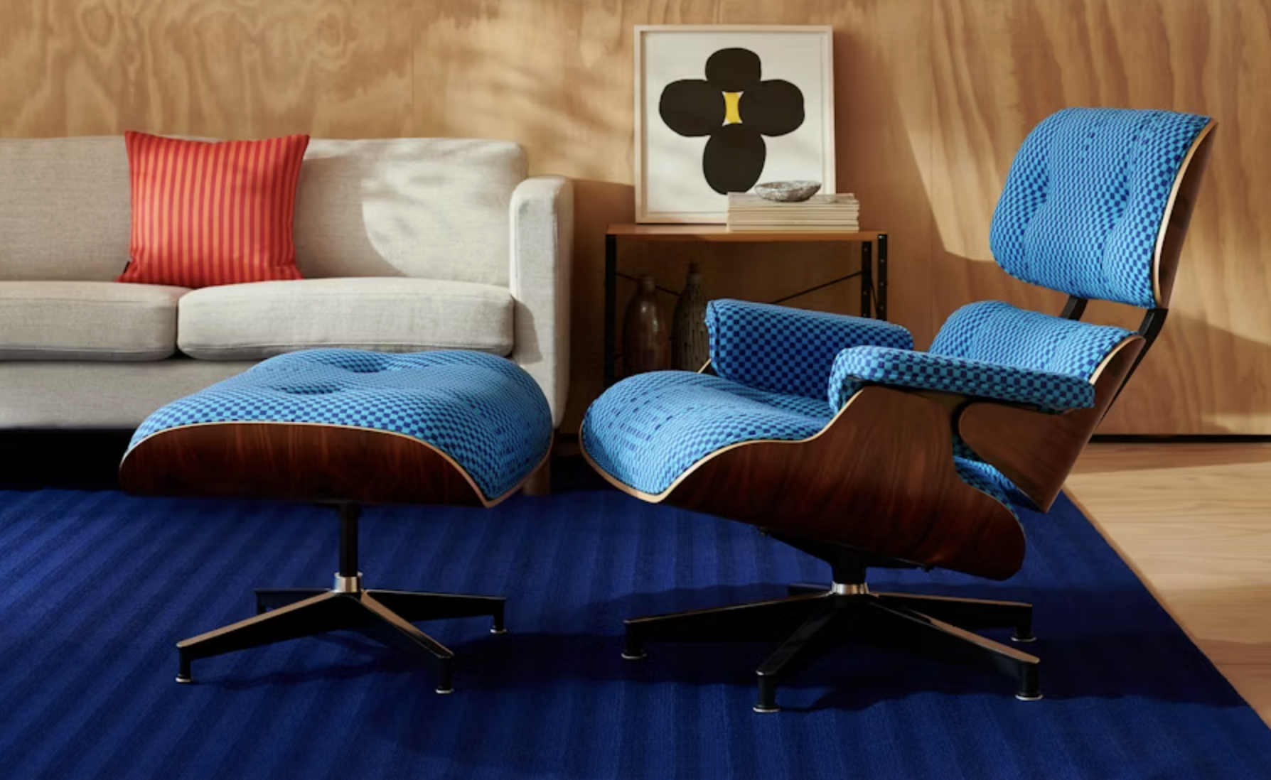 A classic Eames Lounge Chair and Ottoman upholstered in a bright blue and black checkered fabric with dark wood shells sits on a striped blue rug in a modern living space with a light gray sofa and wood-paneled wall.