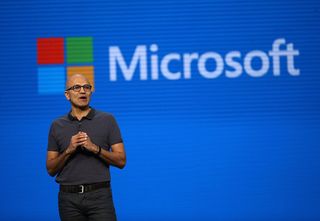 Microsoft CEO Satya Nadella standing in front of a screen displaying the Microsoft logo.