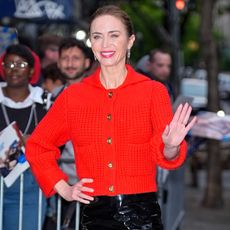 emily blunt waves to a crowd while wearing a bright red sweater