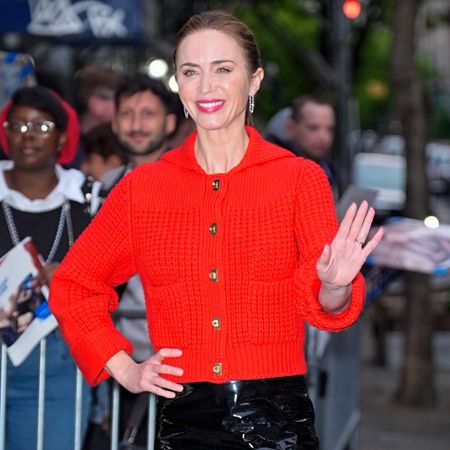 emily blunt waves to a crowd while wearing a bright red sweater