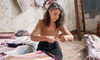 A young woman sits on a wooden floor while cutting an orange thread part of a wider textile sculpture.