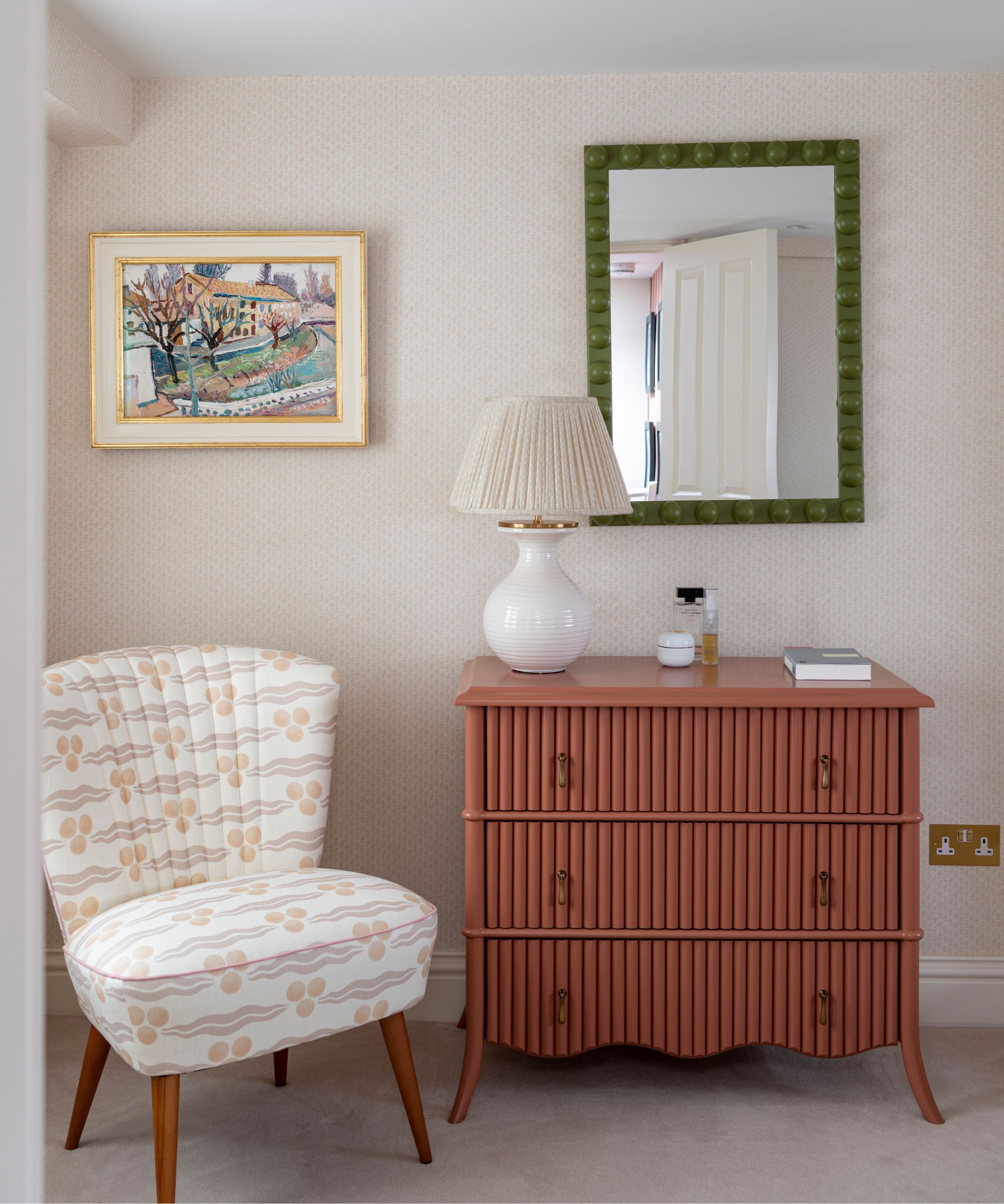A neutral bedroom space with a patterned upholstered chair, brown fluted bedroom drawers with a white lamp, perfumes and book on top, and a green framed mirror above next to a colorful framed artwork in a white and gold frame.