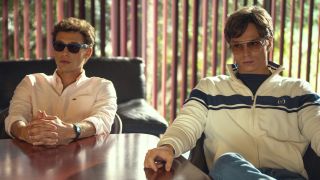 (L to R) Cooper Koch as Erik Menendez, Nicholas Chavez as Lyle Menendez sitting at a table with their hands on it, they're both also wearing sunglasses. 