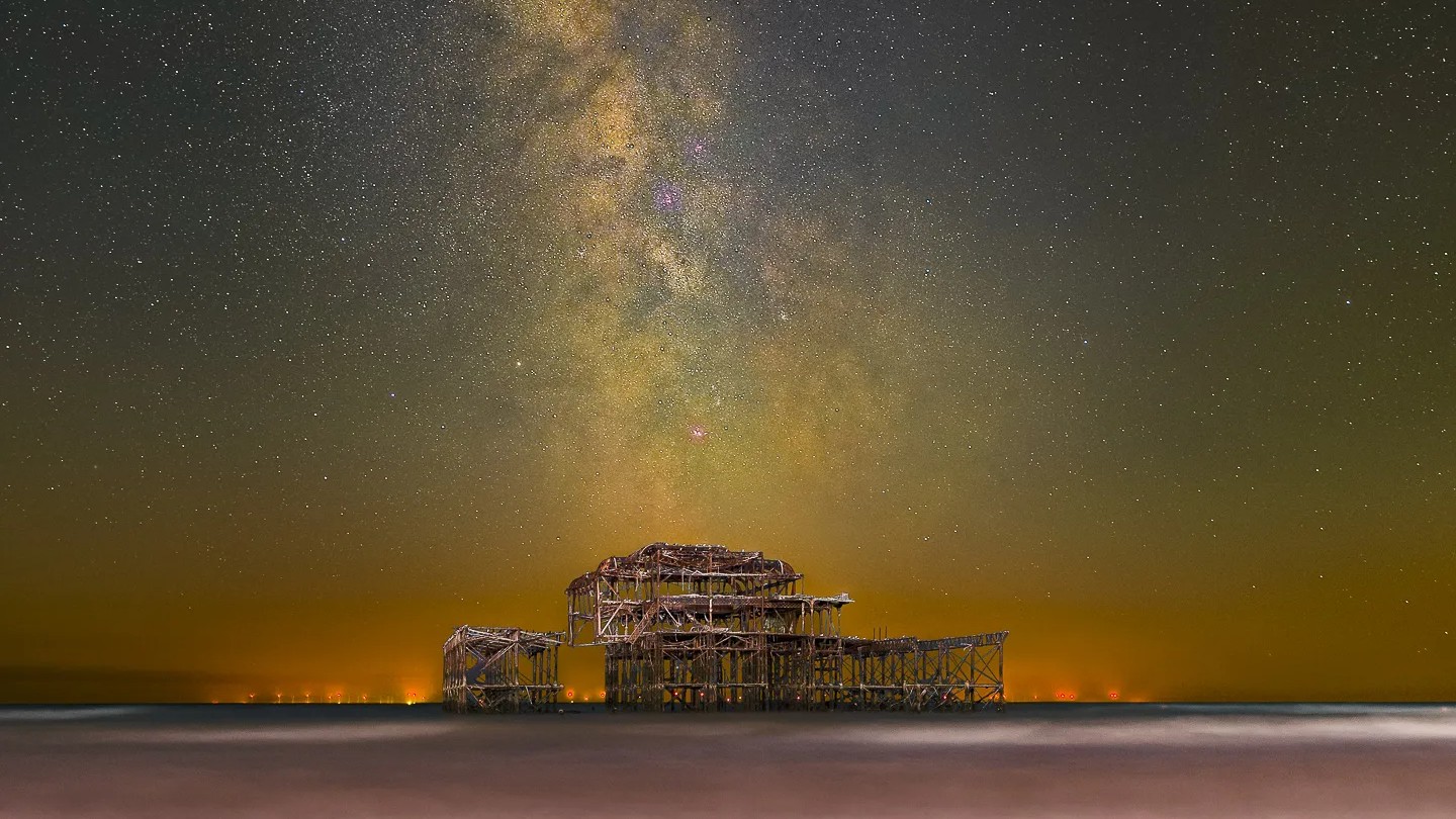 Night photograph of the Milky Way galaxy arcing above the decaying structure of the West Pier in Brighton, with the sea blurred from a long exposure and a pebble beach in the foreground.