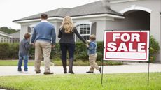 A family of four stands outside a house with a for sale sign.