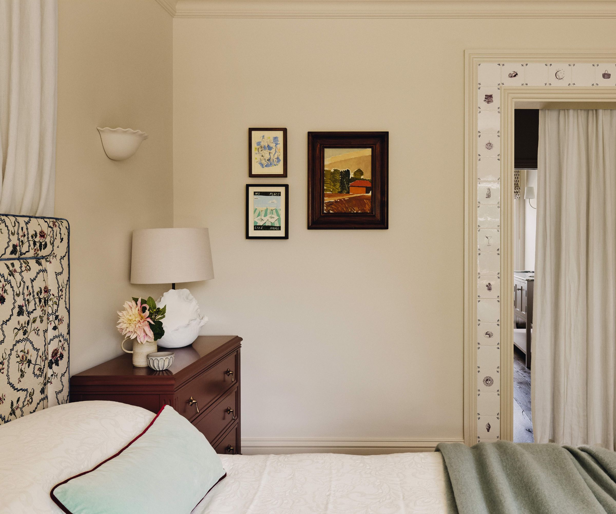 A cream bedroom with an upholstered headboard with a floral pattern, paired with a wooden nightstand. On the wall, there are three small framed artworks. There is a door way through to the bathroom covered with a cream curtain that is surrounded by Delft tiles.