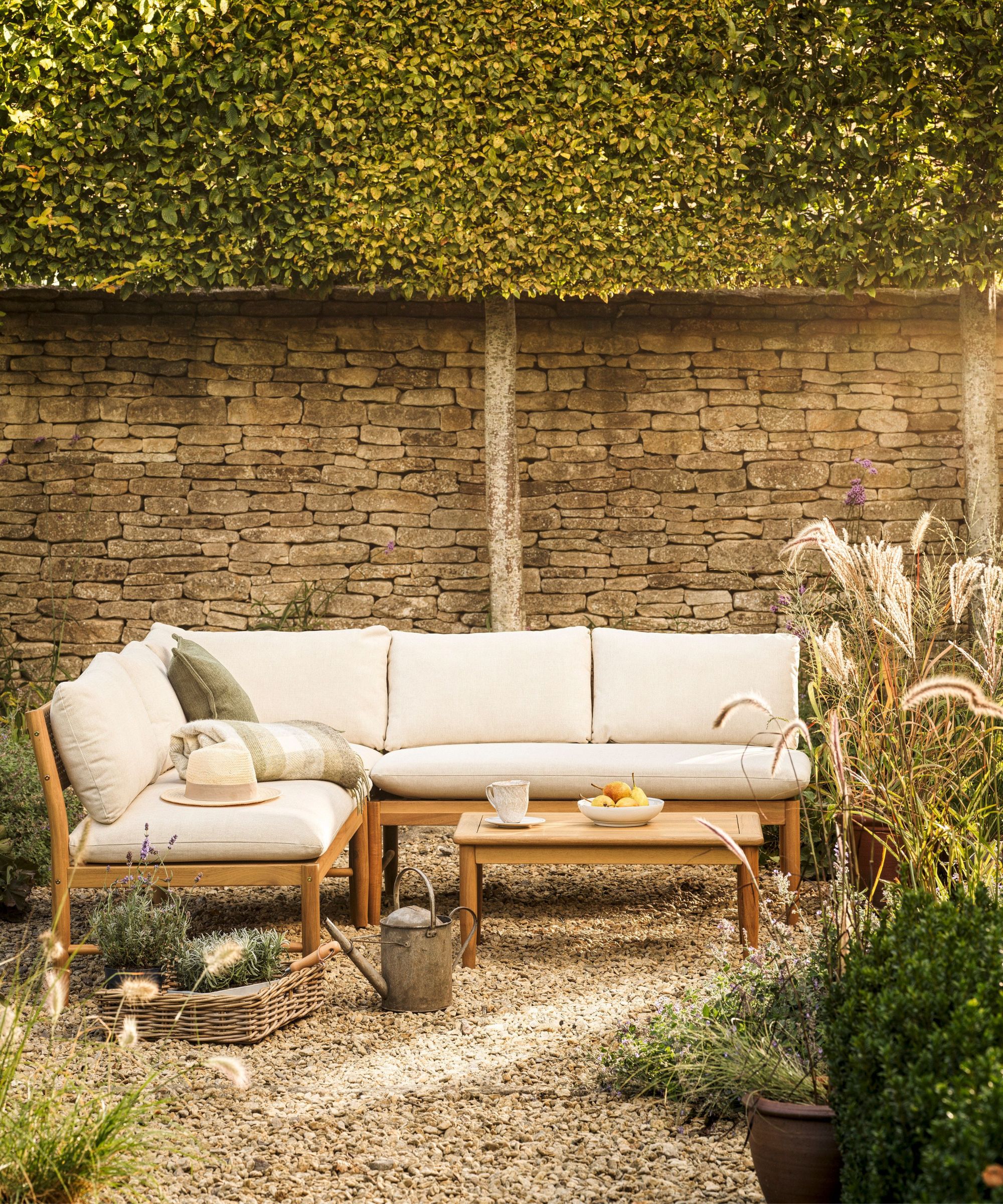 A teak corner sofa with cream cushions and a low teak coffee table on a gravel garden, backed by a Cotswold stone wall with pleached hornbeam hedging above