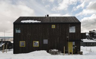 Alternative exterior view of the Oslo family house by STA under a blue cloudy sky - the house features a dark wood exterior and windows and doors with yellow frames. There is snow on the ground and other houses nearby