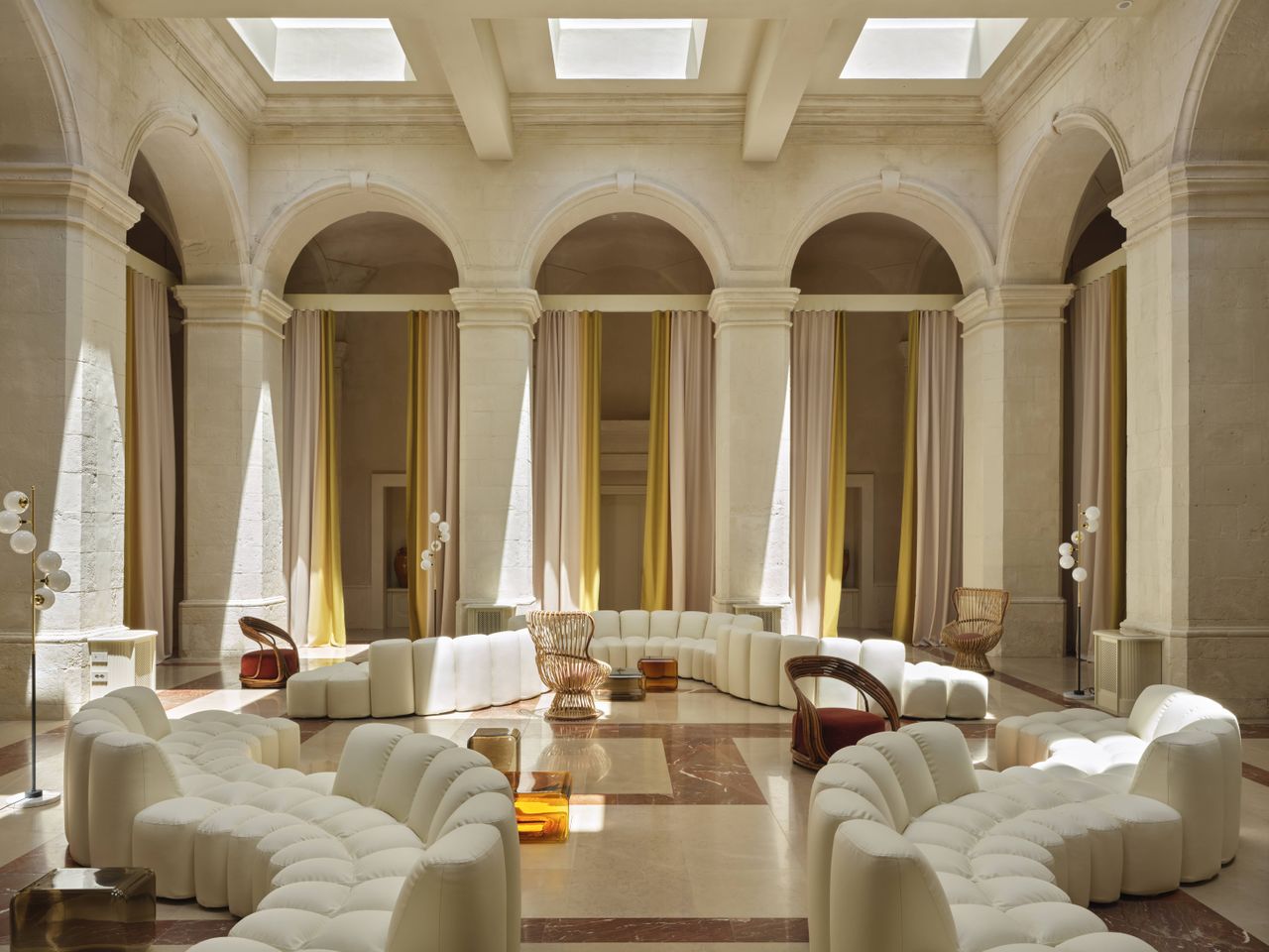A design hotel with historic, vaulted architectural walls houses a selection of retro-inspired furniture, including colorful sculptural armchairs, layered wallpapers and panels, tiled flooring, eye-catching artworks, and wavy sofas, along with a panoramic rooftop bar with sun loungers.