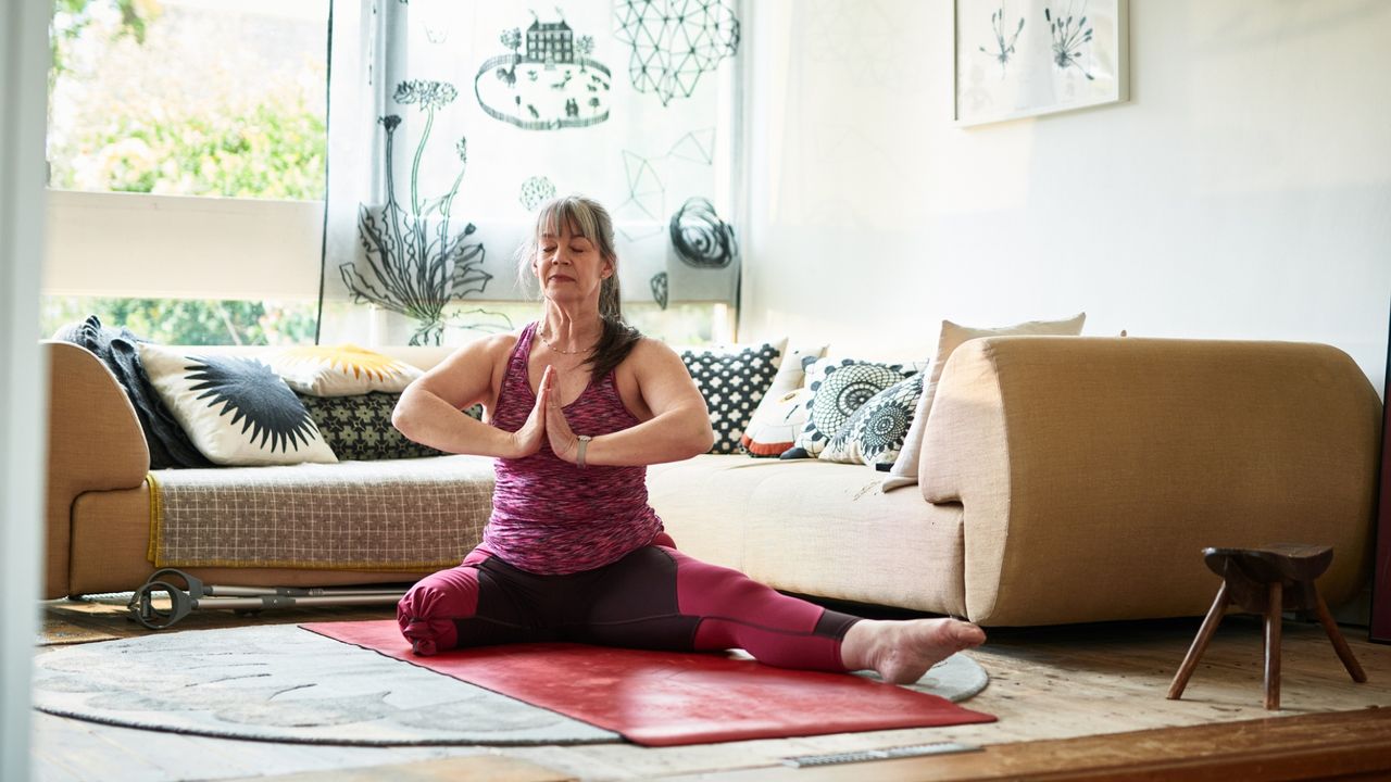 one legged older woman wearing pink vest and leggings sitting on a yoga mat in front of a sofa in a living room setting. her palms are together in a prayer position in front of her chest. 