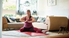 one legged woman sitting on a yoga mat in front of a sofa in a living room setting. her palms are together in a prayer position in front of her chest. 