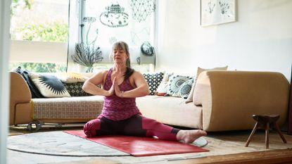 one legged woman sitting on a yoga mat in front of a sofa in a living room setting. her palms are together in a prayer position in front of her chest. 