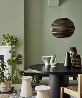 A matt green dining room with a round black dining table, wooden chairs and a low hanging black pendant light