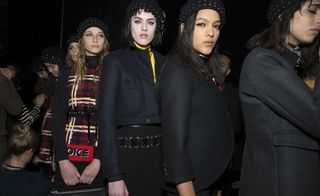 Models backstage wearing black jackets, skirts, red checkered dresses and headwear