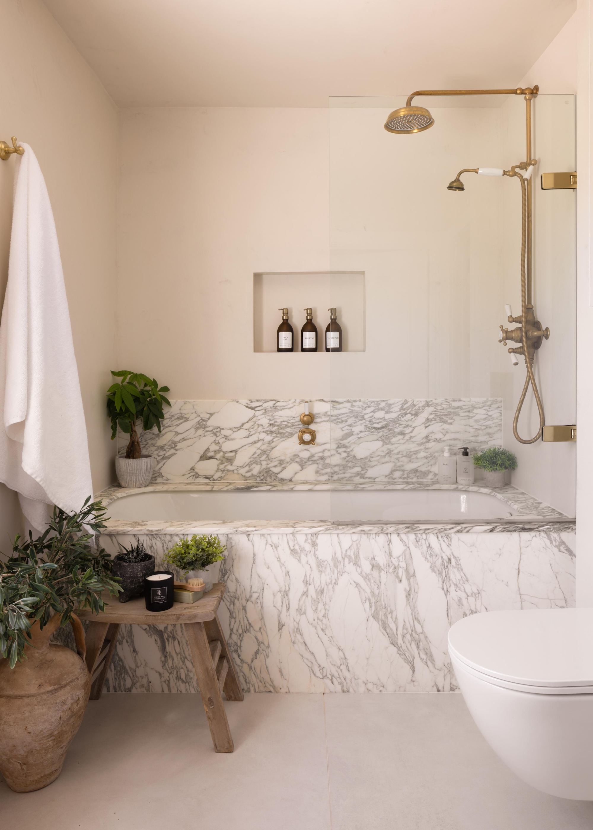 a modern classic bathroom with a marble clad built-in bath and an over bath shower in gold shown with lots of plants, a neat row of toiletries and white towels