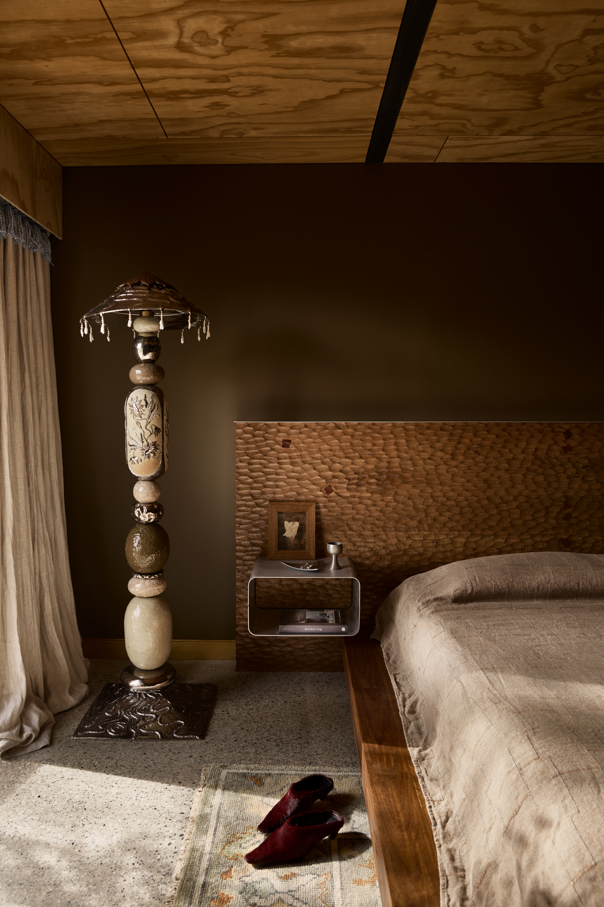 modern bedroom with brown walls, timber walls, chiselled timber bedhead, brown bedcover, sculptural lamp, concrete floors, rug, and pair of shoes