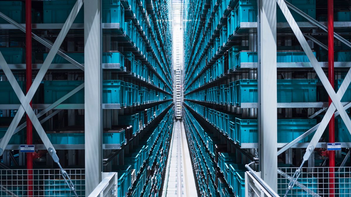 A symmetrical shot of a warehouse full of shelves, with a row of shelves on either side of the frame full of blue crates representing the supply chain.