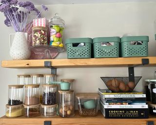 Two wooden shelves in a pantry. The bottom shelf shows stacked glass jars with bamboo lids, recipe books and a wire basket with eggs. The top shelf has decorative purple flowers in white vase, and three sage green plastic storage bins