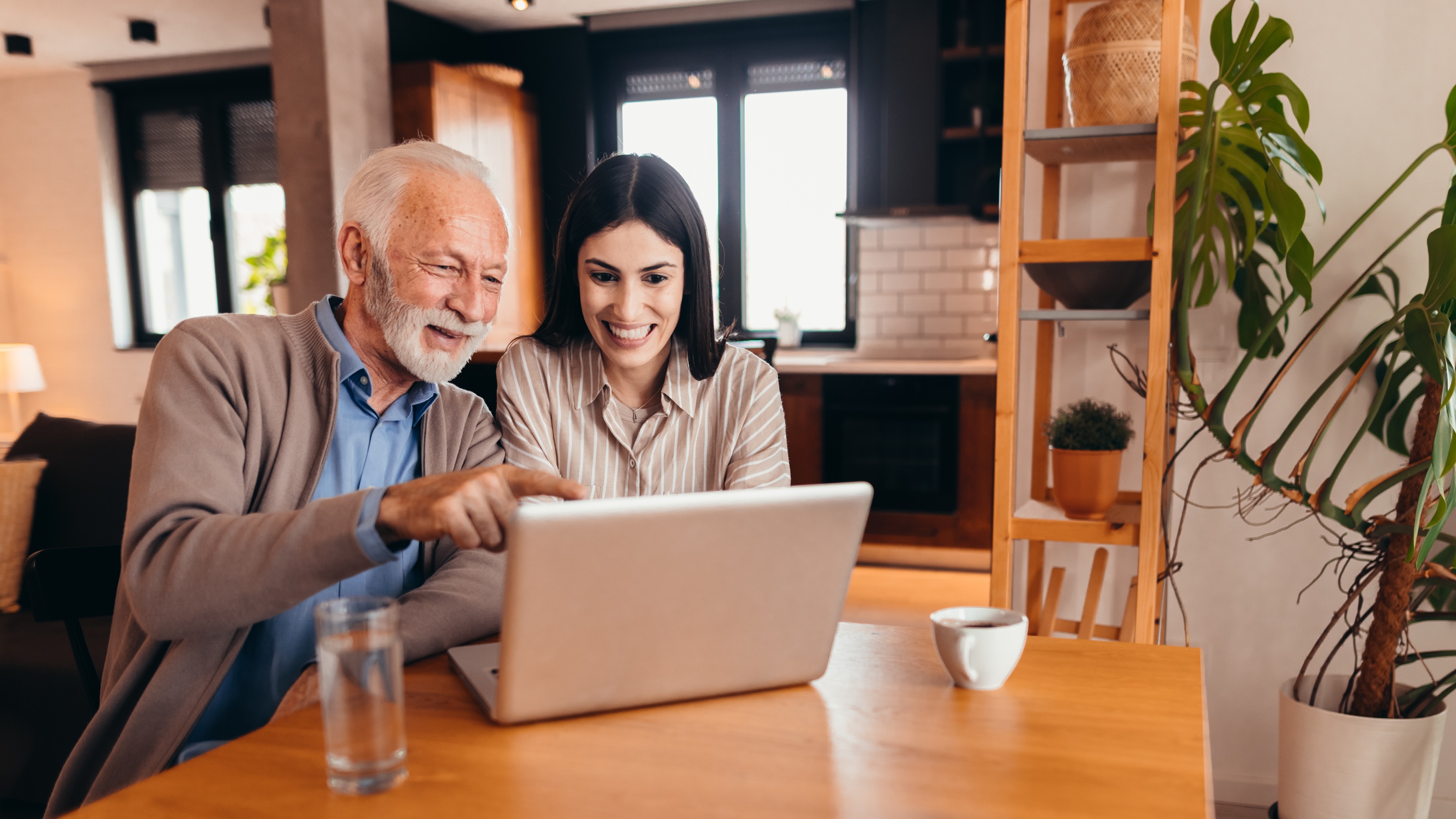 A father smiles while pointing at a laptop while sitting next to his adult daughter at a table.