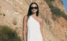 Woman with dark hair and sunglasses wearing white one-shoulder dress standing in desert background