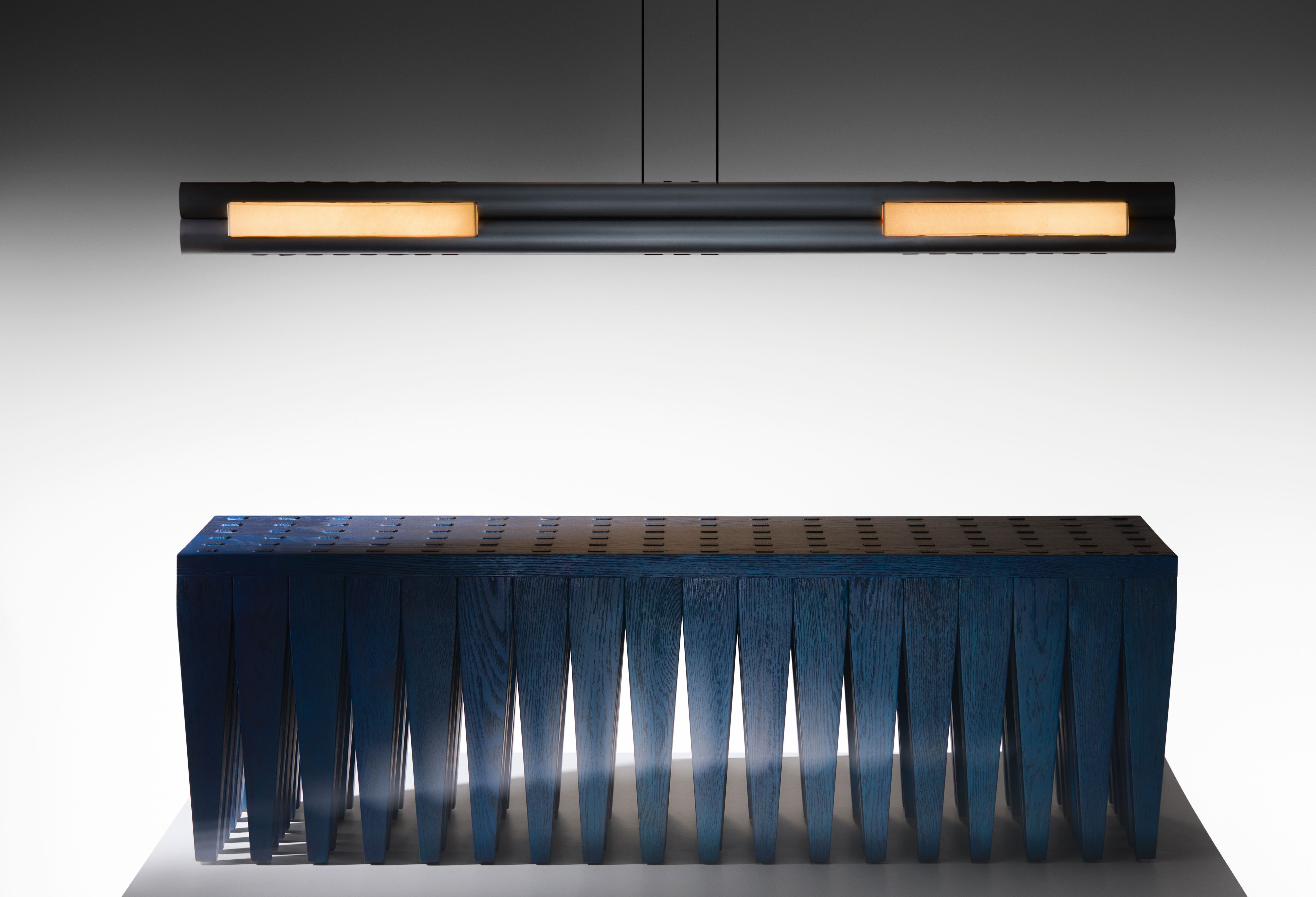 A modern lighting fixture designed by Astraeus Clarke, featuring two laser-cut metal tubes with a calfskin parchment shade, displayed above a textured blue wooden console.
