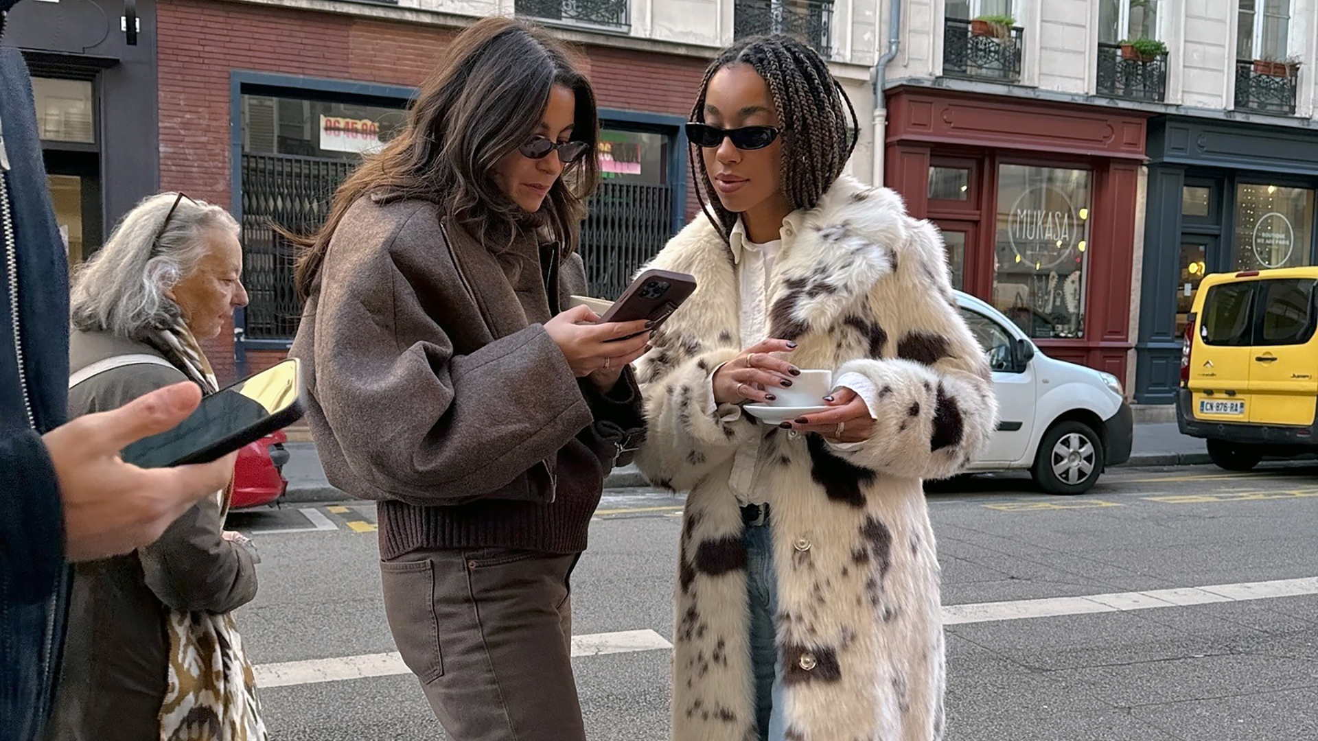 street style shot of Dutch fashion influencer Amaka Hamelijnck wearing black sunglasses and an animal-print coat looking at a phone with a friend that is wearing a brown bomber jacket and jeans