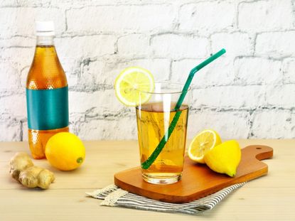 A glass of ginger ale with a green straw and a slice of lemon, surrounded by lemons, a ginger root, and an unlabeled plastic bottle