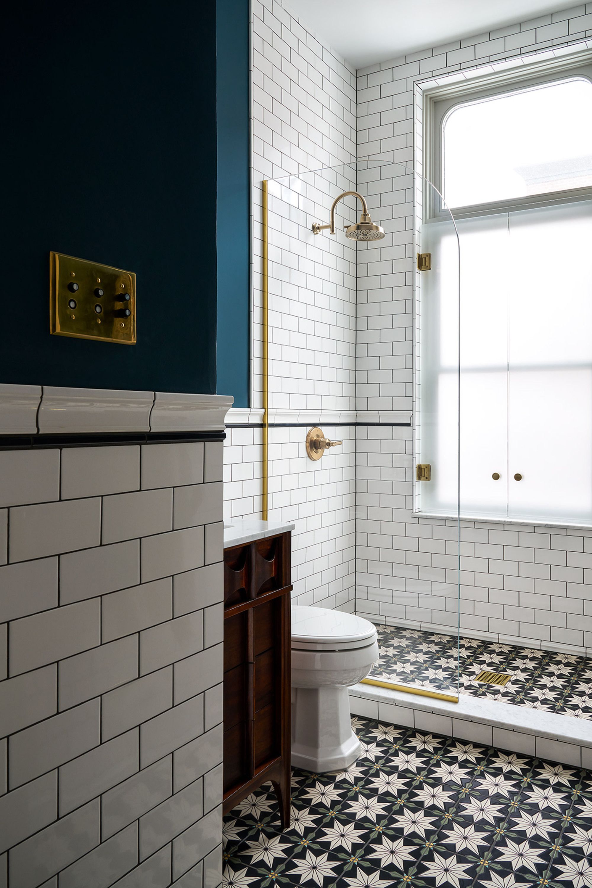 The bathroom is light with white tiles and black grout, the floor is a 70s style pattern of flowers with black, gold and green. The wall above is a dark teal colour 