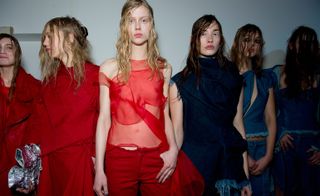 Row of six young female models, three with red outfits to the left, three with denim outfits to the right, long straggly hair style, eye make up, white wall backdrop