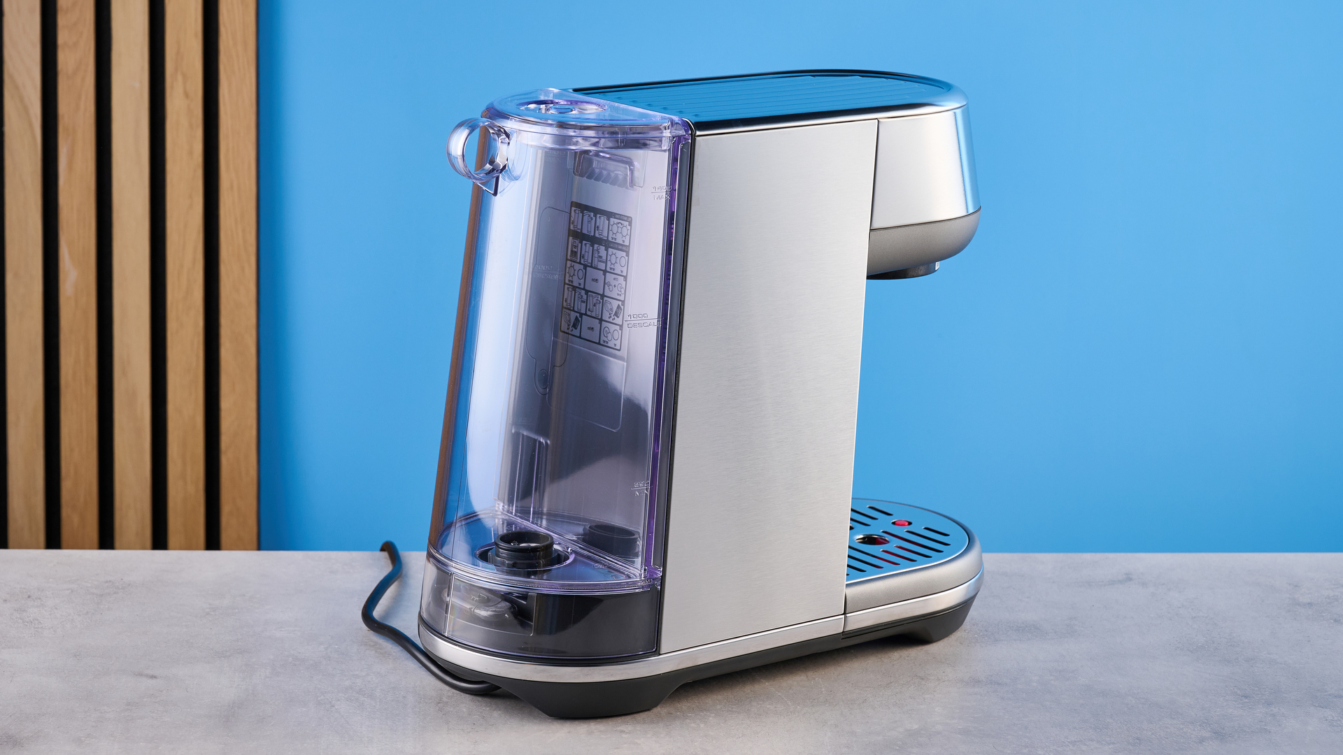 the breville bambino / sage bambino in silver photographed against a blue background with a wooden feature wall, showing the steam wand, group head, water tank, button controls, and espresso