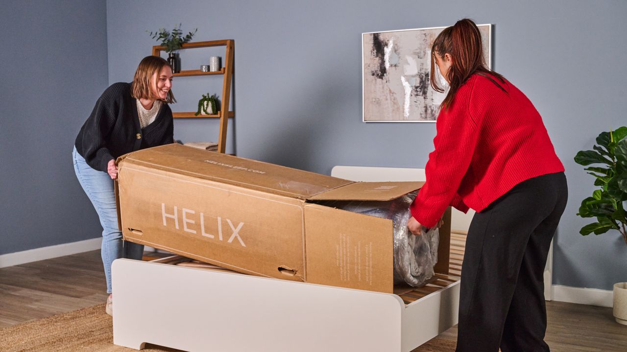 The image shows two women taking a Helix Midnight mattress out of its box on a white bed frame