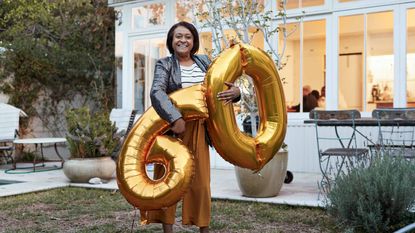 A smiling woman holds two big balloons in the shape of a 6 and a 0.